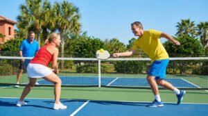 Three smiling individuals play pickleball on a vibrant outdoor court. Man in yellow hits ball, woman in red awaits, man in blue watches. Sunny, palm trees.